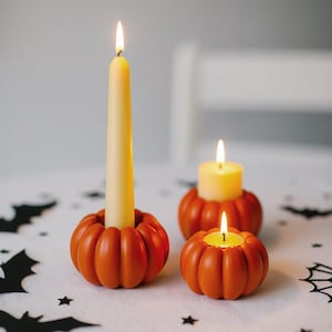 Peut inclure: Trois bougeoirs orange en forme de citrouille avec des bougies allumées. Une grande bougie crème est dans un support, tandis que deux petits supports contiennent des bougies jaunes allumées. Les supports sont sur une surface blanche avec des décorations d'Halloween.