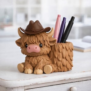 May include: A brown Highland cow-shaped desk organizer with a cowboy hat. The pen holder has a shaggy fur design and holds a pink, purple, and black pen. The cow has a pink nose and is sitting on a white surface.