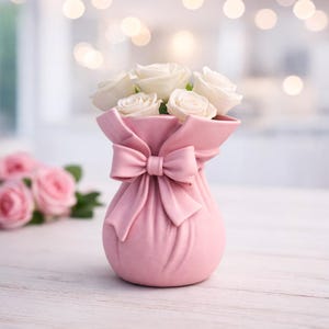 Peut inclure: Un vase rose en céramique en forme de sac avec un nœud, contenant des roses blanches. Le vase a une surface texturée et est posé sur une table en bois blanche. Arrière-plan flou avec des lumières bokeh.
