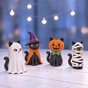 May include: Four small cat figurines dressed in Halloween costumes. One cat wears a white ghost sheet, one wears a purple witch hat and orange cape, one wears a carved pumpkin head with a green bow tie, and one is wrapped in white mummy bandages.