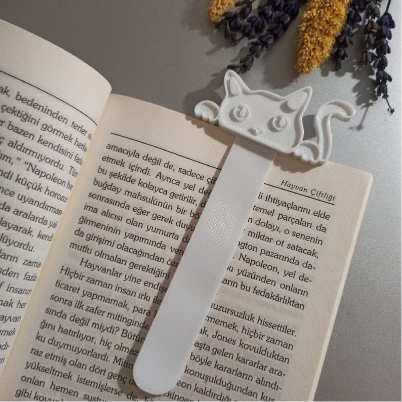 以下が含まれることがあります： かわいい漫画の顔をした猫の形をした、白の3Dプリントブックマーク。ブックマークは、トルコ語のテキストが書かれた開いた本のの上にあります。ブックマークのテキストは「Hayvan &Ccedil;iftliği」です。