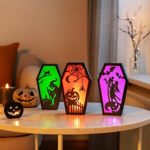 May include: Three coffin-shaped Halloween lanterns with colourful interior lights. Each lantern features a different spooky design: a witch, a spider web, and a silhouette of a couple with bats. The lanterns are on a round table.