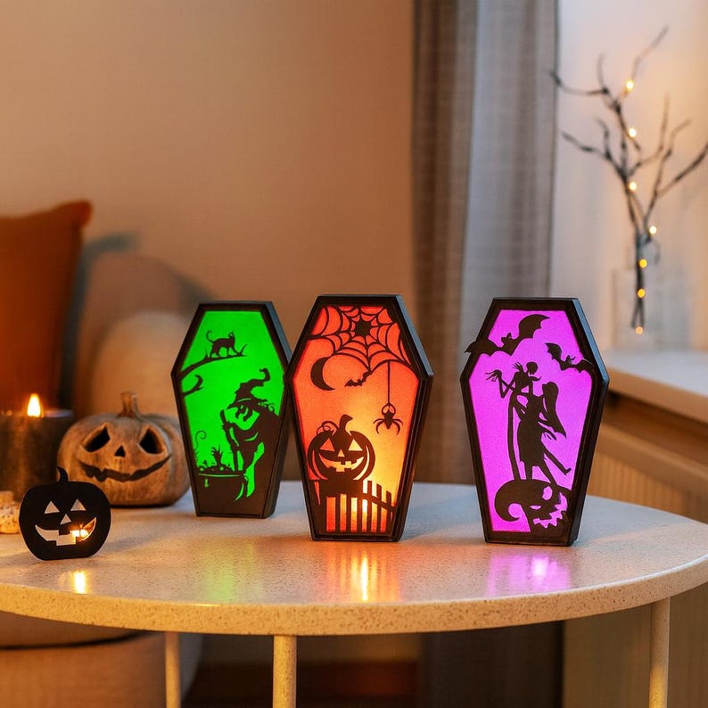 May include: Three coffin-shaped Halloween lanterns with colourful interior lights. Each lantern features a different spooky design: a witch, a spider web, and a silhouette of a couple with bats. The lanterns are on a round table.