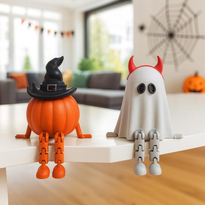 May include: Two Halloween-themed figurines sit on a white surface. One is an orange pumpkin wearing a black witch hat. The other is a white ghost with red horns. Both have articulated legs and are designed to sit on a ledge.