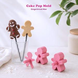 Peut inclure: Un moule à cake pop rose en forme de bonhomme en pain d'épices, accompagné de deux cake pops finis sur des bâtonnets dans un verre transparent. Le texte "Cake Pop Mold" et "Gingerbread Man" est imprimé en haut de l'image.