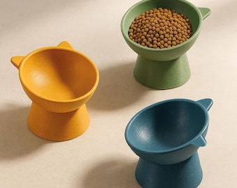Puede incluir: Tres cuencos para comida de mascotas con forma de gato en amarillo, verde y azul. Los cuencos están elevados sobre un soporte y tienen un diseño de oreja de gato en el borde.