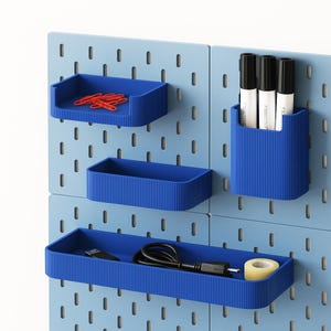 May include: A light blue pegboard wall organizer system featuring four modular dark blue storage attachments. The attachments include a shallow tray with red paperclips, a tall pen holder with three black markers, a small rectangular bin, and a long tray holding a black USB cable and a roll of tape.