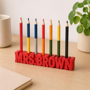 Puede incluir: Un organizador de escritorio rojo con el texto MRS. BROWN en letras de bloque, diseñado con siete ranuras verticales que sostienen lápices de colores. Los lápices tienen cuerpos de dos tonos en rojo, amarillo, azul, naranja, verde y negro.