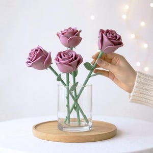 Könnte beinhalten: Ein klares Glasvase enthält vier malvenfarbene Rosen mit grünen Stielen und Blättern. Eine Rose wird von einer Hand gehalten. Die Vase steht auf einem Holztablett, vor einem weißen Hintergrund mit sanfter Beleuchtung.