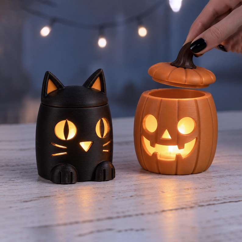 Pu&ograve; includere: Un contenitore nero a forma di gatto con occhi e naso gialli luminosi, posizionato accanto a un contenitore arancione a forma di zucca con un volto intagliato e un coperchio rimovibile, entrambi illuminati dall'interno da una luce calda.