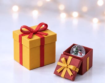 May include: A yellow square gift box with a red ribbon and bow, alongside an open red gift box with a yellow ribbon and bow containing a small glass snow globe featuring a miniature green Christmas tree and a snowman with a black top hat.