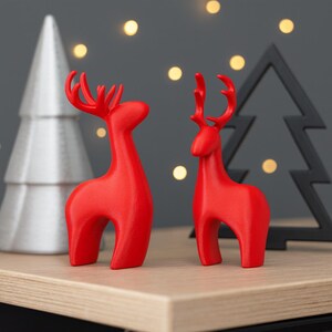 May include: Two red reindeer figurines with stylized antlers stand on a wooden surface. A silver Christmas tree and a black tree-shaped decoration are in the background, with soft, out-of-focus lights.
