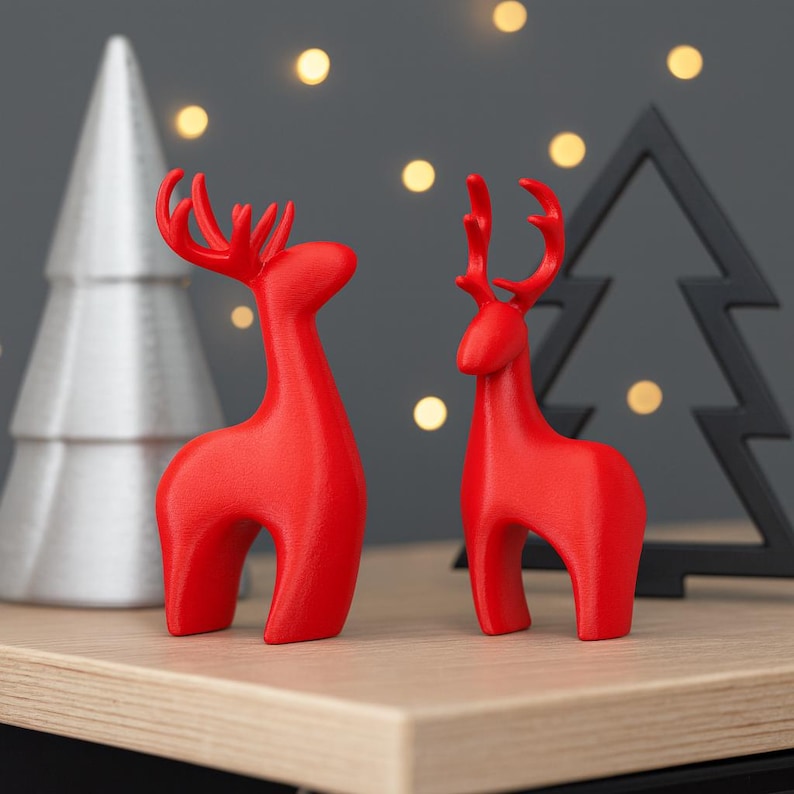 May include: Two red reindeer figurines with stylized antlers stand on a wooden surface. A silver Christmas tree and a black tree-shaped decoration are in the background, with soft, out-of-focus lights.