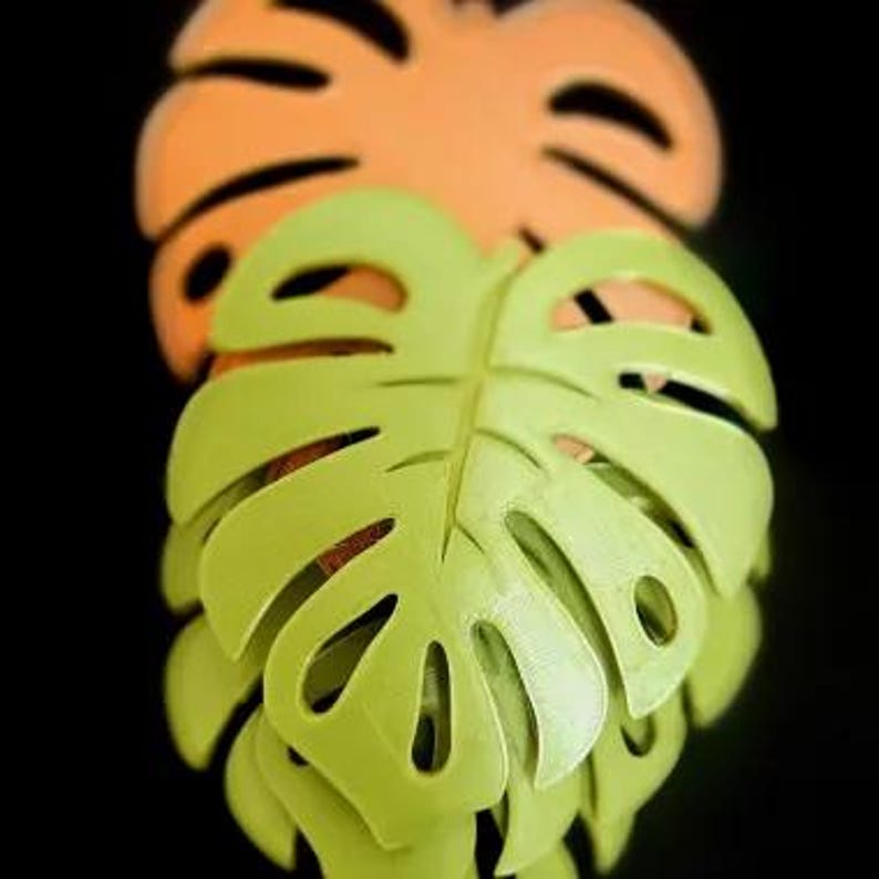Puede incluir: Dos recortes decorativos de hojas de Monstera. La hoja delantera es verde claro, y la hoja de atr&aacute;s es naranja. Las hojas tienen una apariencia tridimensional y est&aacute;n sobre un fondo negro.