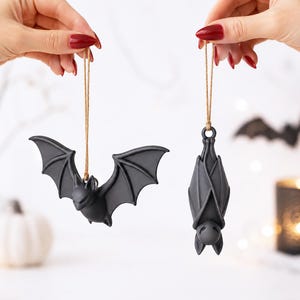 May include: Two matte black bat ornaments hanging from thin brown twine strings. One bat is depicted with its wings fully spread in a flying position, while the other bat is shown in a vertical, resting position with its wings folded tightly inward.