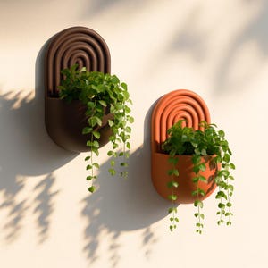 Op de afbeelding: Twee wandplantenbakken in bruin- en oranjetinten, elk met een halfronde bovenkant met concentrische bogen. Groene planten vallen uit de plantenbakken en vormen een contrast met de neutrale muur. Ontworpen voor binnen of buiten.