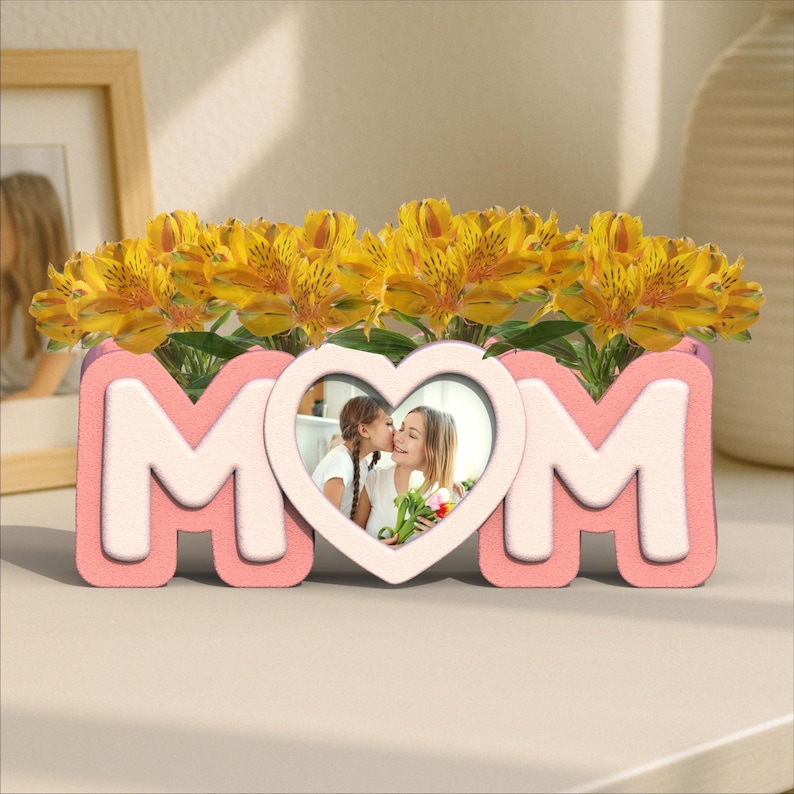 K&ouml;nnte beinhalten: Ein rosa-wei&szlig;er Keramik-Pflanztopf in Form des Wortes "MOM" mit einem herzf&ouml;rmigen Bilderrahmen in der Mitte. Der Topf ist mit gelben Blumen und einem Foto einer Mutter und ihrer Tochter gef&uuml;llt.