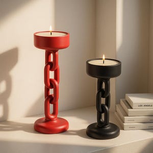 May include: Two modern candle holders. One is red, the other black. Each has a round base, a chain-link style stem, and a circular top holding a lit candle. The candles are white, and the flames are visible. The candle holders are on a white surface.