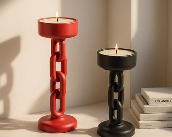 Puede incluir: Dos candelabros modernos, uno rojo y otro negro. Cada uno tiene una base redonda, un tallo estilo eslabón de cadena y una parte superior circular que sostiene una vela encendida. Las velas son blancas y las llamas son visibles. Los candelabros están sobre una superficie blanca.
