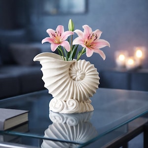Może przedstawiać: Kremowy wazon w kształcie muszli trzyma różowe lilie z zielonymi łodygami i pąkami. Wazon stoi na szklanym stole, odbijając wazon i kwiaty. Tło jest rozmyte i ciemnoniebieskie.