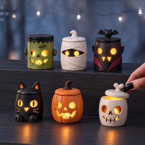Pu&ograve; includere: Una collezione di sei piccoli portacandele cilindrici a tema Halloween, raffiguranti un mostro di Frankenstein verde, una mummia bianca, un vampiro nero con un pipistrello, un gatto nero, una zucca arancione e un teschio bianco con coperchio.