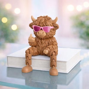 May include: A brown figurine of a Highland cow wearing pink sunglasses and giving the middle finger. The cow is sitting on a white book with articulated legs. The figurine has a shaggy coat and small horns.