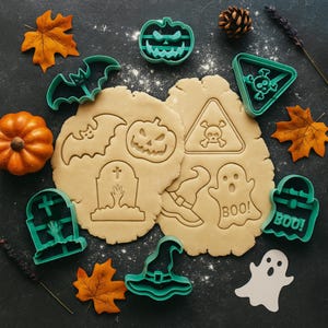 Puede incluir: Cortadores de galletas con temática de Halloween en color verde azulado, que incluyen una calabaza, un murciélago, un sombrero de bruja, una lápida y un fantasma. La imagen también muestra galletas cortadas con diseños a juego, una pequeña calabaza y hojas de otoño.