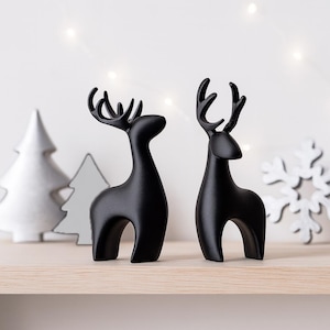 May include: Two black reindeer ornaments with detailed antlers stand on a wooden shelf. A white Christmas tree and snowflake are in the background, with soft lights adding a festive touch. The reindeer are a modern holiday decoration.