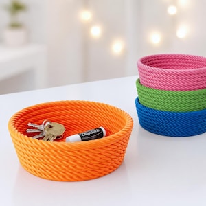 May include: A set of four round, rope-coiled storage bowls in bright orange, pink, green, and blue. The orange bowl contains a metal key ring with several keys and a white ChapStick lip balm tube with black text. The bowls are stacked and open.