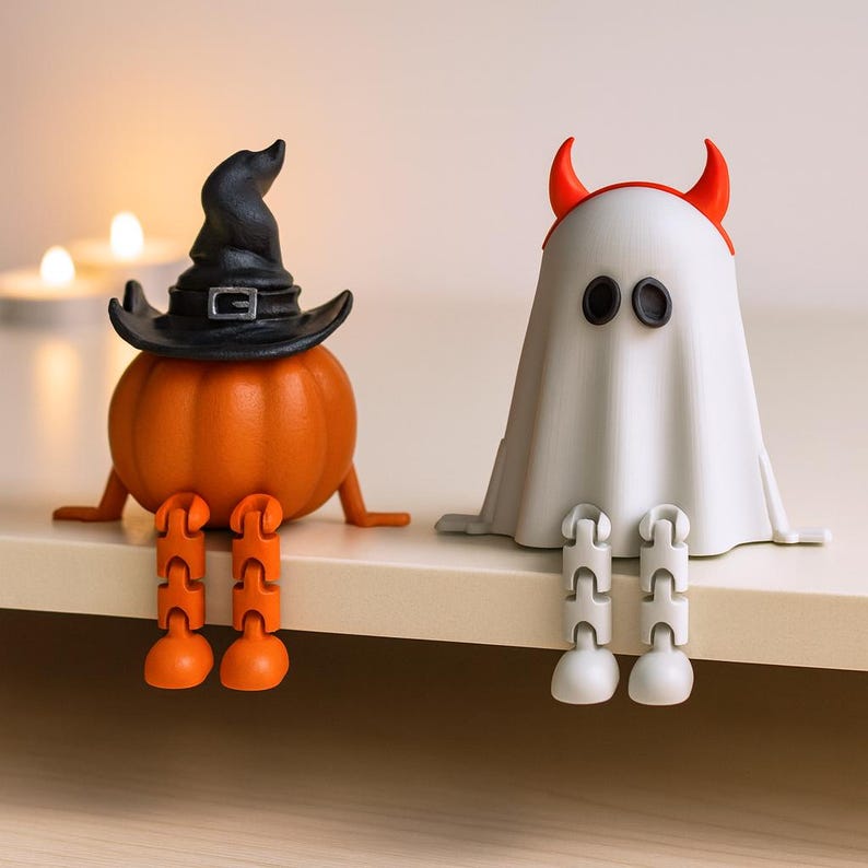 May include: Two Halloween-themed figurines sit side-by-side. One is an orange pumpkin wearing a black witch hat. The other is a white ghost with red horns. Both have jointed legs and are sitting down.