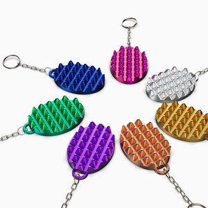 May include: A collection of colorful keychains in a variety of metallic finishes. Each keychain features a textured, oval-shaped design with numerous raised, triangular spikes. The keychains are arranged in a circular pattern.
