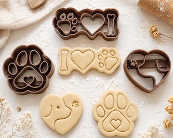 May include: A set of brown plastic cookie cutters and matching dough cutouts featuring dog-themed designs. The shapes include a paw print, a heart with a dog profile, and a rectangular plaque with a bone, heart, and paw print motif.