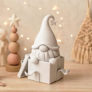 May include: A white gnome figurine emerging from a gift box. The gnome has a long, curved hat with a star on the tip and a long, fluffy beard. The box has a ribbon-like detail. A wooden star-topped tree is in the background.