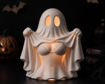 May include: A matte white ceramic ghost figurine illuminated from within, featuring two oval eye cutouts and a draped sheet design that reveals a bikini top and midriff. The ghost is positioned upright with its arms raised, holding the fabric edges.