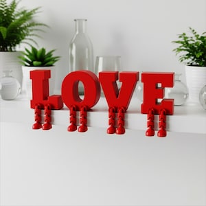 May include: Red "LOVE" letters with articulated legs, sitting on a white shelf. The letters are a vibrant red, with each letter having a pair of small, jointed legs. Decorative plants and glass bottles are in the background.