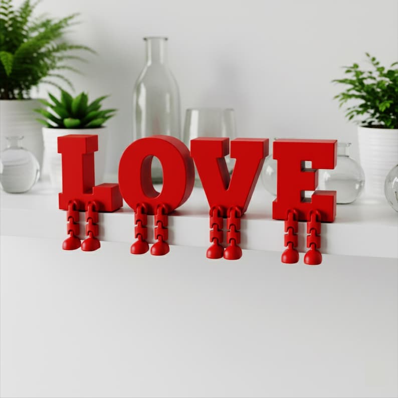 May include: Red "LOVE" letters with articulated legs, sitting on a white shelf. The letters are a vibrant red, with each letter having a pair of small, jointed legs. Decorative plants and glass bottles are in the background.