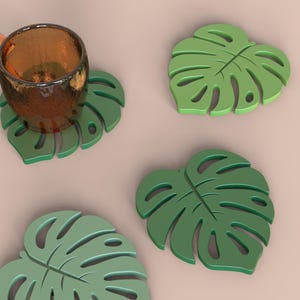 Puede incluir: Un juego de cuatro posavasos verdes con forma de hoja. Uno de los posavasos sostiene un vaso &aacute;mbar texturizado. Los posavasos est&aacute;n dise&ntilde;ados para parecerse a hojas de Monstera, con un color verde claro y detalles intrincados.
