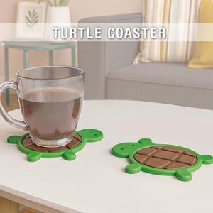 Op de afbeelding: Twee groene schildpadvormige onderzetters met bruine centra liggen op een witte tafel. Een heldere glazen mok gevuld met een donkere drank rust op een onderzetter. De tekst "TURTLE COASTER" wordt hierboven weergegeven.