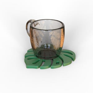 Puede incluir: Una taza de vidrio transparente con una superficie texturizada se encuentra sobre un posavasos verde en forma de hoja. La taza tiene un asa y una abertura ancha. El posavasos es de color verde vibrante.