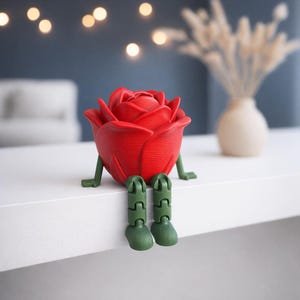 Puede incluir: Un objeto decorativo en forma de rosa roja, con piernas y pies verdes, sentado sobre una superficie blanca. La rosa tiene pétalos detallados y un diseño caprichoso. El fondo incluye un jarrón borroso con flores secas e iluminación suave.