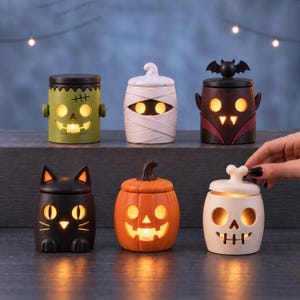 Könnte beinhalten: Ein Set aus sechs Halloween-Teelichthaltern aus Keramik, bestehend aus einem grünen Frankenstein-Monster, einer weißen Mumie, einem schwarzen Vampir mit Fledermausflügeln, einer schwarzen Katze, einem orangefarbenen Kürbis und einem weißen Totenkopf.