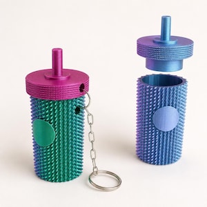 May include: Two cylindrical containers with textured, spiked surfaces. One is teal with a magenta lid and a metal keychain attached. The other is blue with a matching blue lid that is detached and hovering above the open container body.