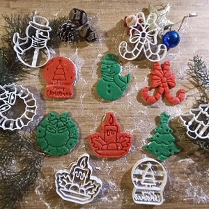 Op de afbeelding: Een verzameling kerst-thema koekjessnijders en koekjes. De snijders zijn van wit plastic en de koekjes zijn rood en groen, in de vorm van sneeuwmannen, kerstbomen, kransen en sneeuwbollen. De koekjes hebben de tekst "Merry Christmas".