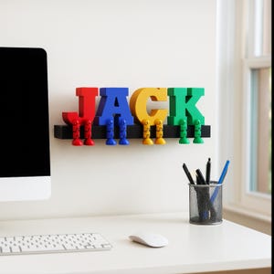 May include: A colorful wall-mounted name sign spelling "JACK". The letters are red, blue, yellow, and green, each with matching legs. The sign is mounted on a black shelf, perfect for a child's room or playroom.
