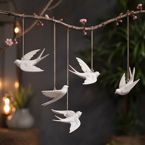 May include: Five white ceramic swallow ornaments hanging from a branch with pink blossoms. The birds are suspended by thin, natural-colored cords, creating a delicate and airy display. The swallows are in various poses, with wings spread and in flight.