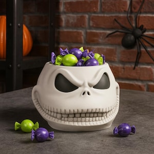 Può includere: Una ciotola di caramelle di Halloween in ceramica bianca a forma di volto di Jack Skellington, piena di caramelle avvolte viola e verdi. La ciotola ha orbite nere e un ampio sorriso. Una decorazione di ragno nero è sullo sfondo.