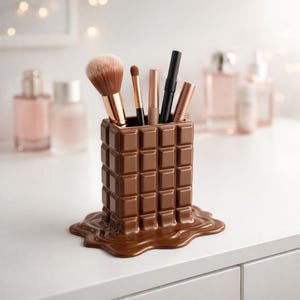 May include: A brown, chocolate bar-shaped makeup brush holder with a melted chocolate base. The holder contains various makeup brushes and applicators. The design is a unique and decorative addition to any vanity or makeup collection.