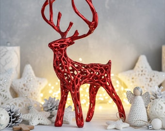 May include: A red decorative reindeer figurine with an open, lattice-like design. The reindeer is standing on a white surface, surrounded by Christmas decorations, including stars, pinecones, and an angel figurine. Warm lights are in the background.