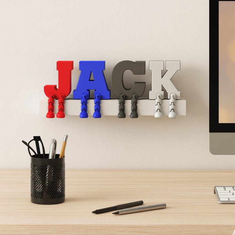 May include: A decorative shelf with the name "JACK" spelled out in colorful, 3D letters. The letters are red, blue, gray, and white, each with attached legs. A black pen holder with pens and pencils sits on a wooden desk.
