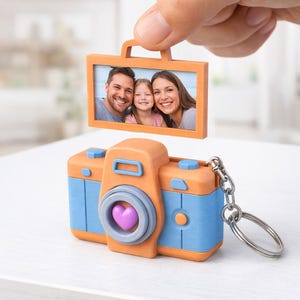Puede incluir: Un llavero novedoso con forma de cámara, con detalles en naranja y azul. La lente de la cámara tiene un corazón morado. Un pequeño marco de fotos con un retrato familiar se sostiene encima. El llavero tiene una cadena y un anillo plateados.